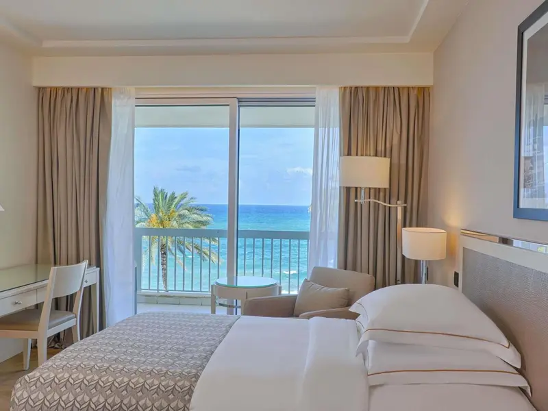 Une chambre lumineuse avec un lit fait et une vue sur la mer depuis un balcon.