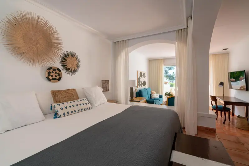 Une chambre lumineuse avec un lit fait et un espace salon visible au fond.