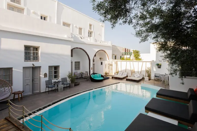 Une piscine rectangulaire turquoise est entourée de chaises longues et de végétation luxuriante devant un bâtiment blanc.