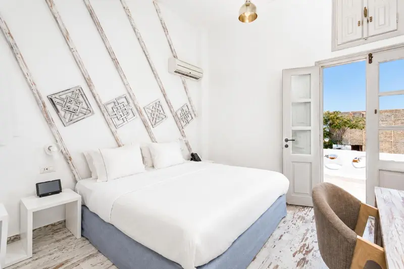 Chambre lumineuse avec un lit recouvert de blanc et une porte-fenêtre ouverte sur une vue extérieure.