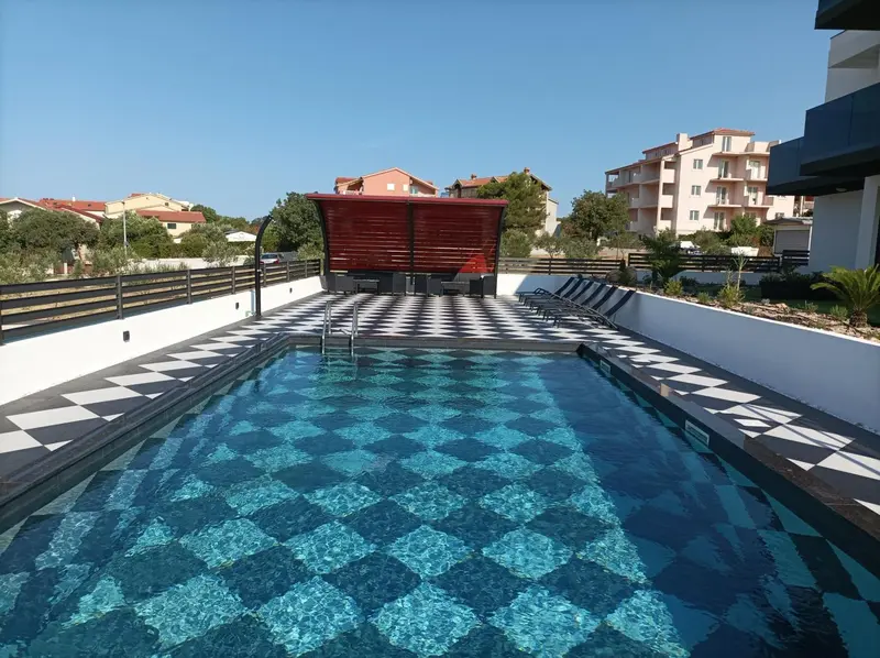 Une piscine rectangulaire aux carreaux bleus et blancs, avec des transats et un espace ombragé.