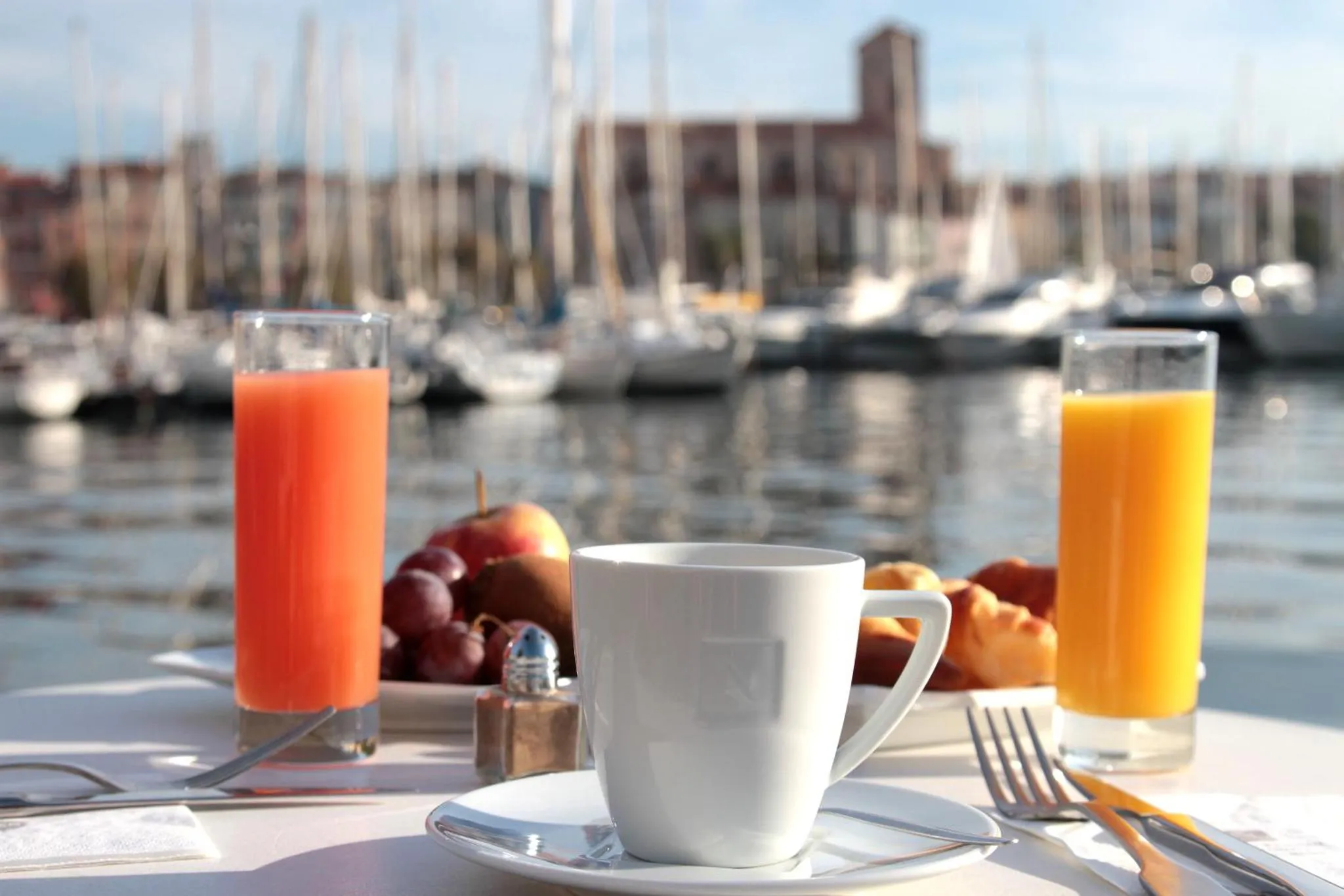 Une table est dressée avec des jus d'orange, des viennoiseries et des fruits, face à un port de plaisance.