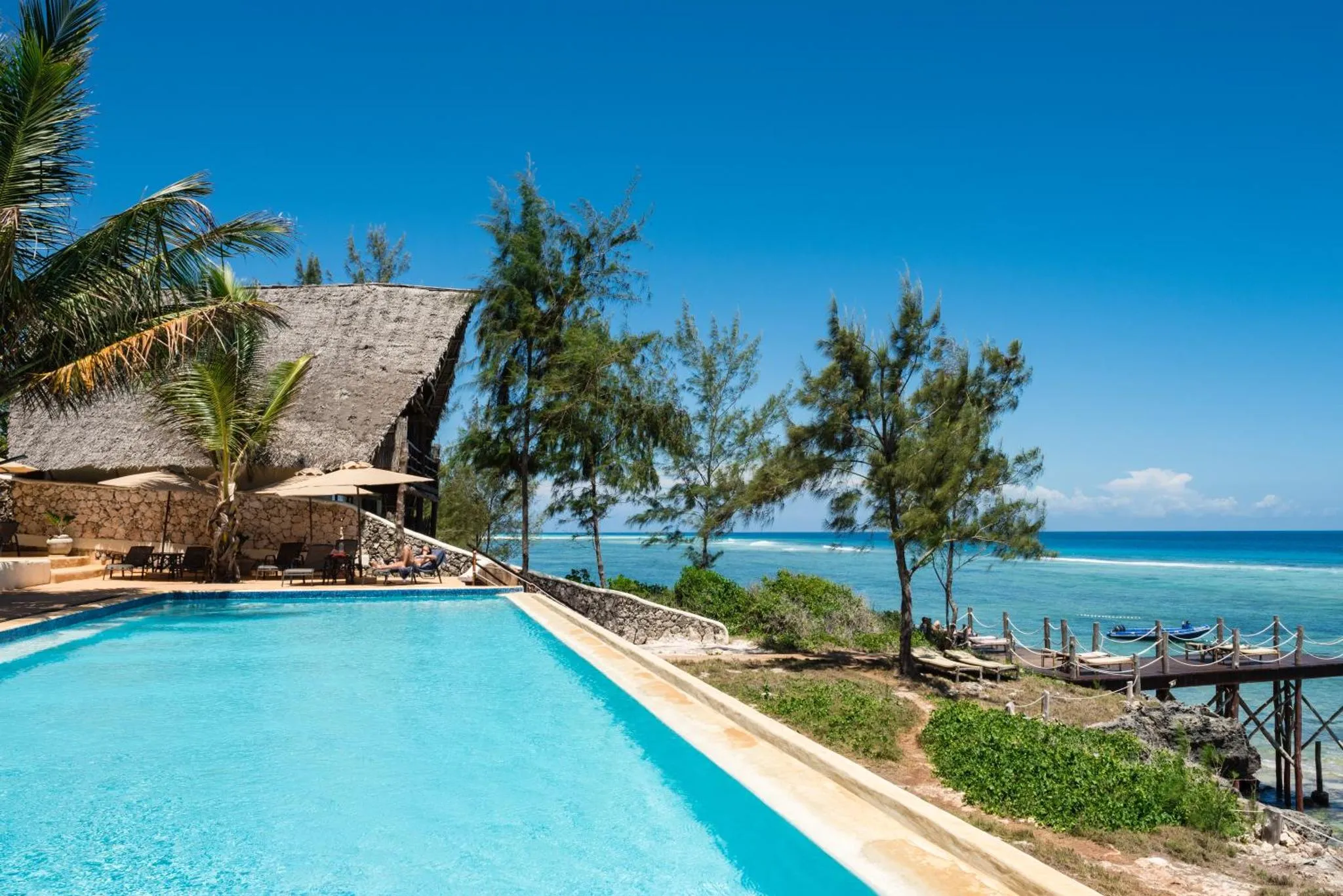 Une piscine bleue bordée de transats, avec vue sur la mer turquoise et une plage.