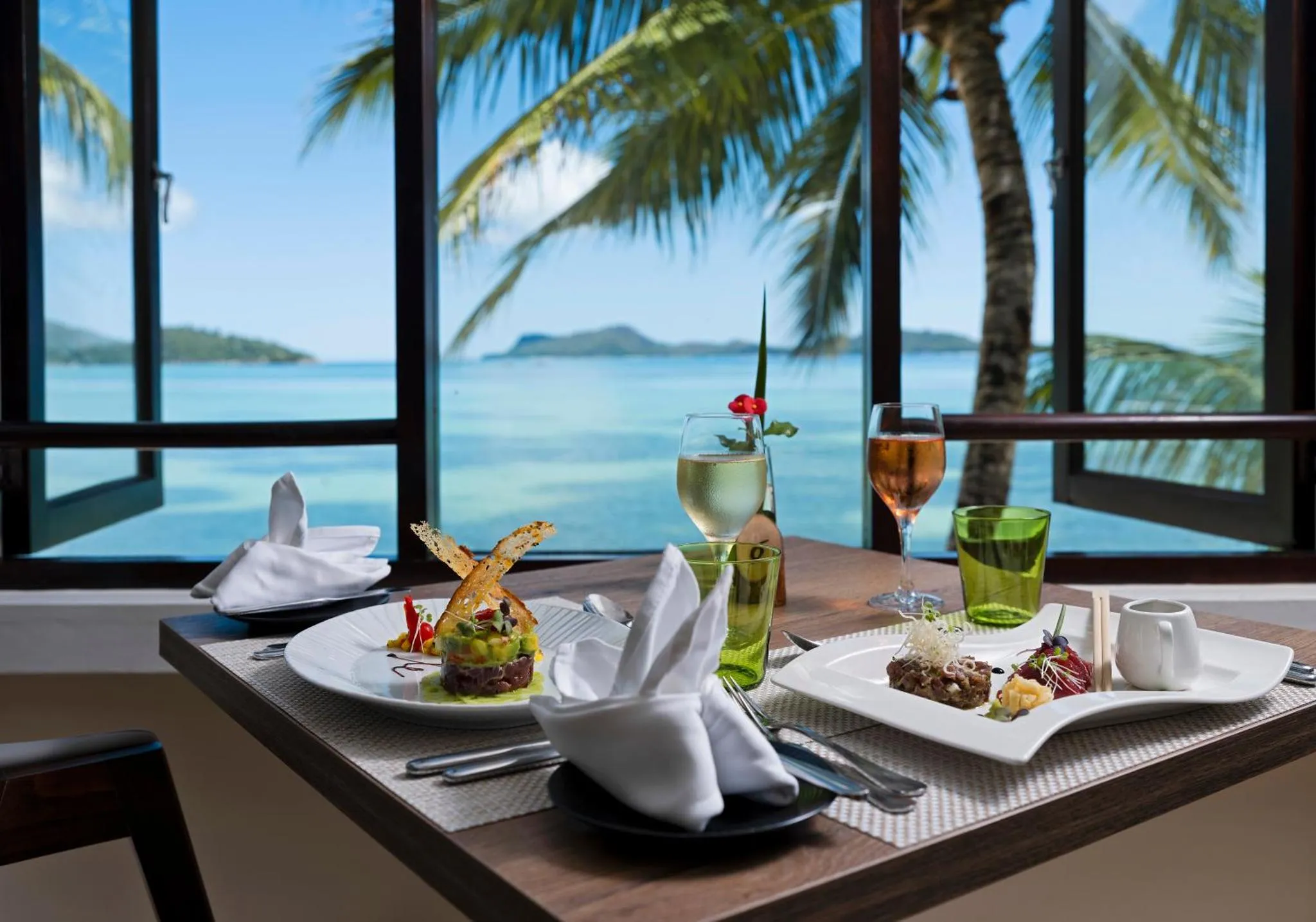 Une table dressée avec des plats et des boissons offre une vue sur la mer et la végétation tropicale.