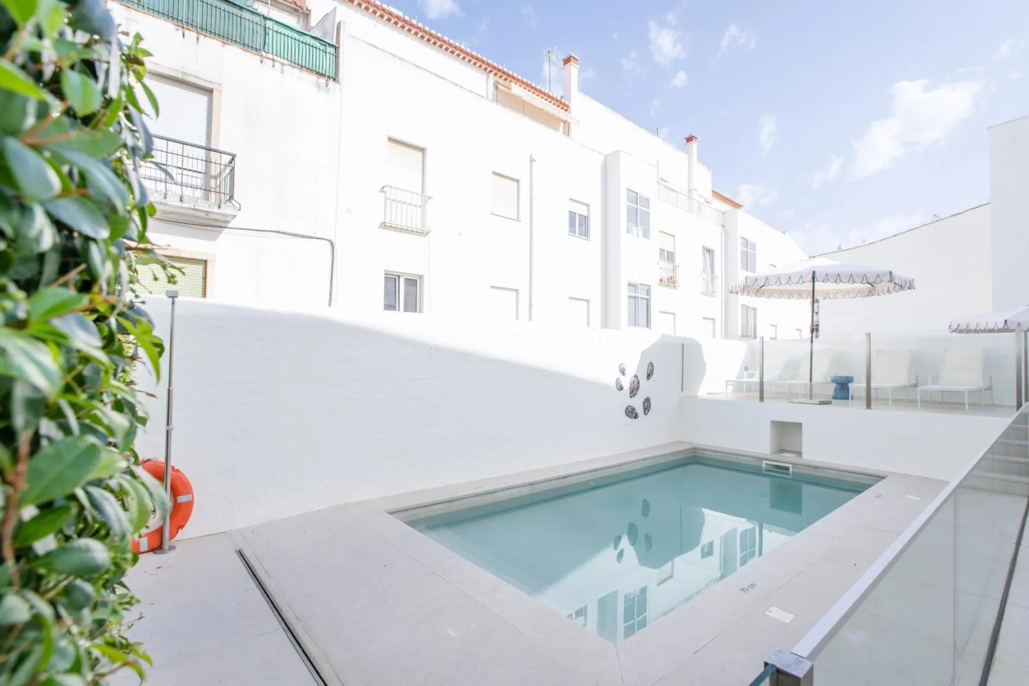 Petite piscine rectangulaire entourée d'une terrasse blanche et de bâtiments clairs.