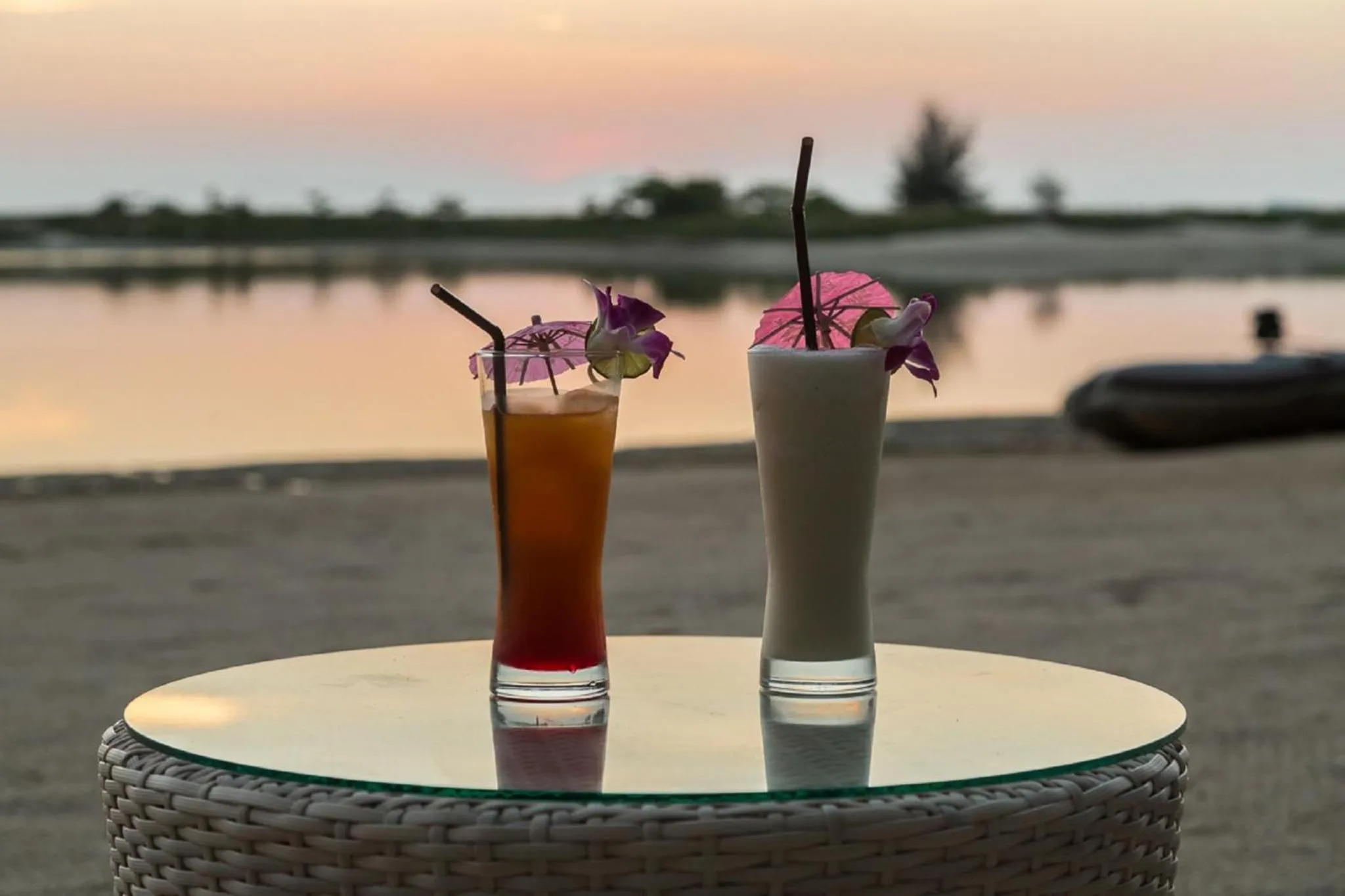 Deux cocktails colorés sont présentés sur une table ronde au bord de l'eau au coucher du soleil.