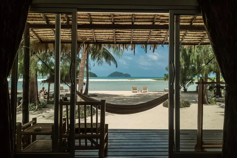 Vue sur une plage de sable blanc et une mer turquoise depuis une terrasse ombragée.