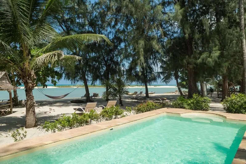 Une piscine turquoise bordée de chaises longues et de végétation tropicale, avec une plage en arrière-plan.