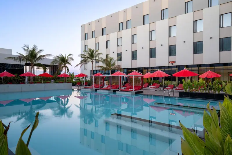 Une piscine turquoise reflète un bâtiment moderne et des palmiers sous un ciel crépusculaire.
