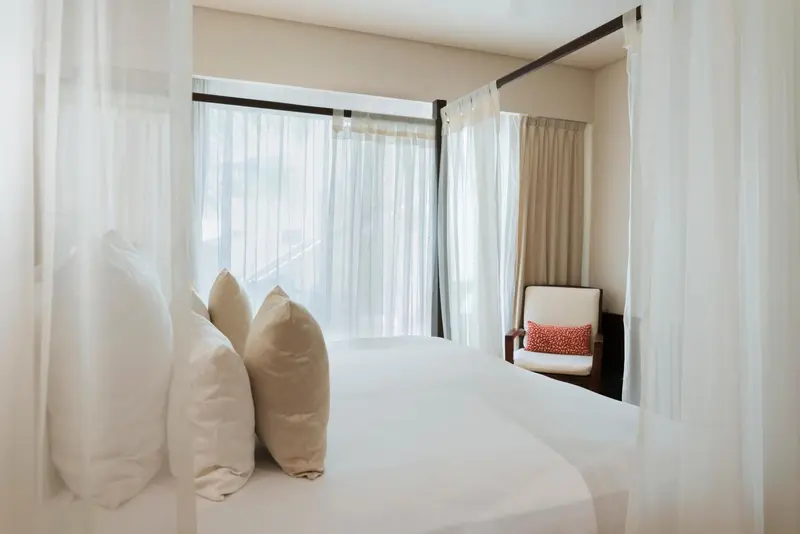 Lit blanc avec coussins et fauteuil beige dans une chambre lumineuse.