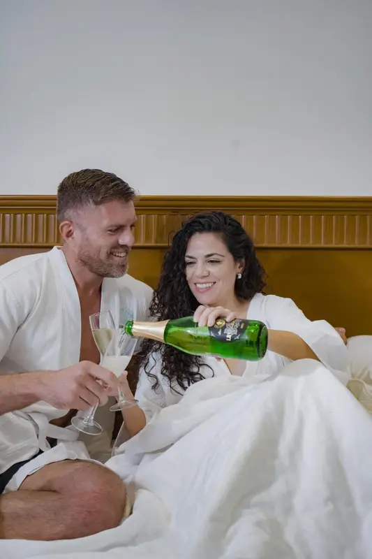Un couple rit en versant du champagne dans des flûtes sur un lit.