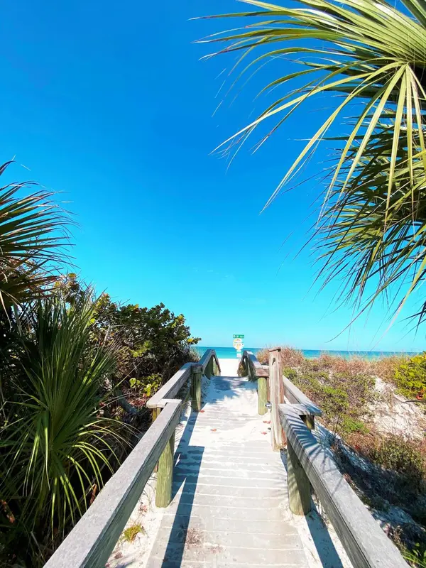 Une passerelle en bois mène vers une plage de sable blanc sous un ciel bleu dégagé.