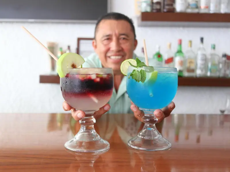 Un barman présente deux cocktails colorés sur un comptoir en bois.