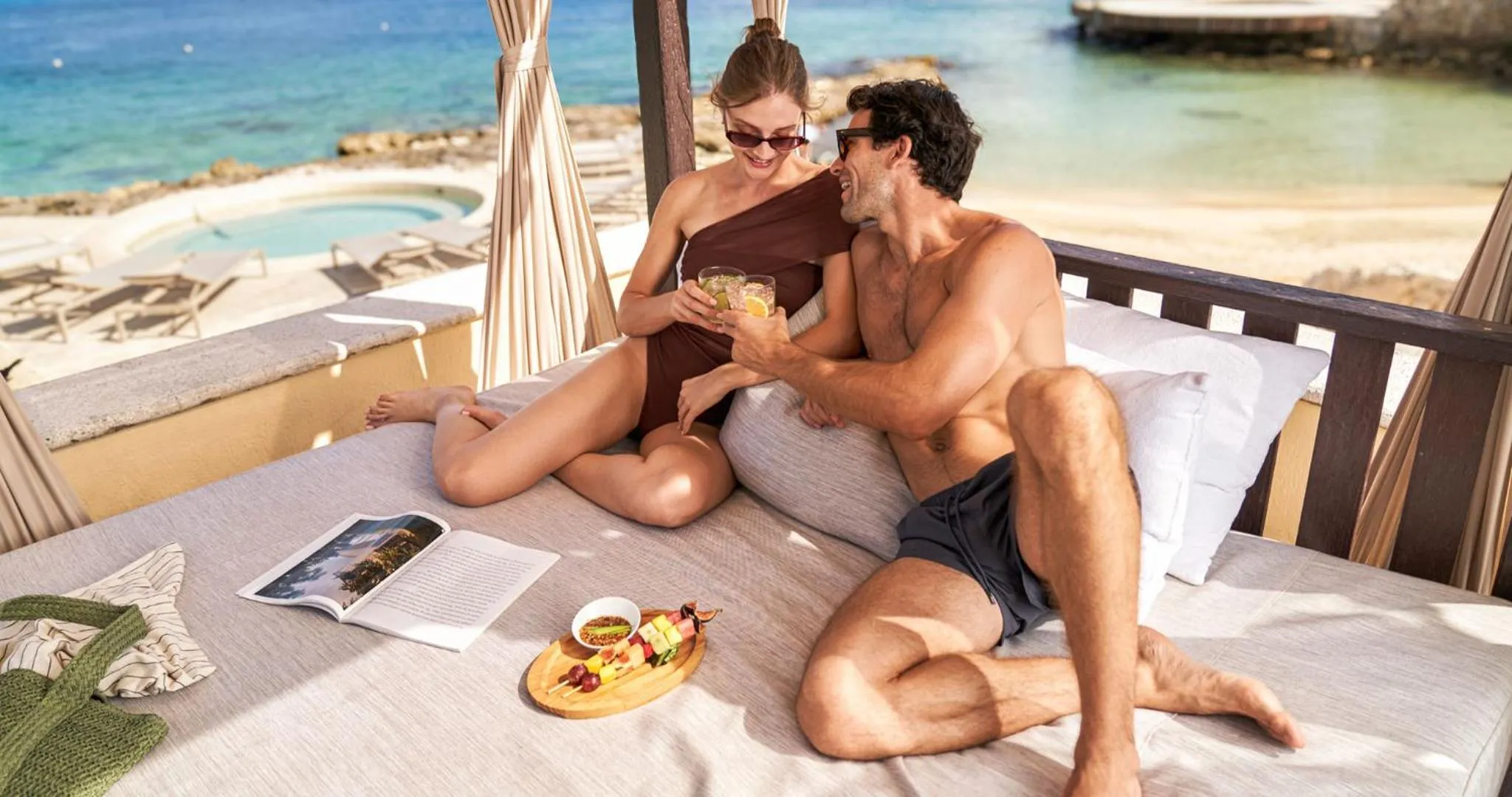 Un couple se détend sur des coussins blancs avec des boissons et des en-cas, face à la mer.
