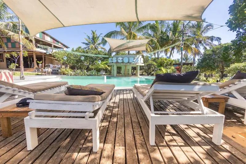 Transats blancs sur une terrasse en bois devant une piscine et un bâtiment tropical.