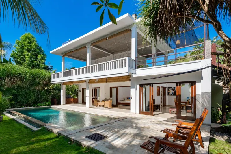 Bâtiment blanc à deux étages avec une piscine et des chaises longues dans un jardin tropical.