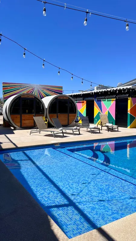 Piscine rectangulaire turquoise bordée d'une terrasse ensoleillée avec des chaises longues et des structures cylindriques colorées.