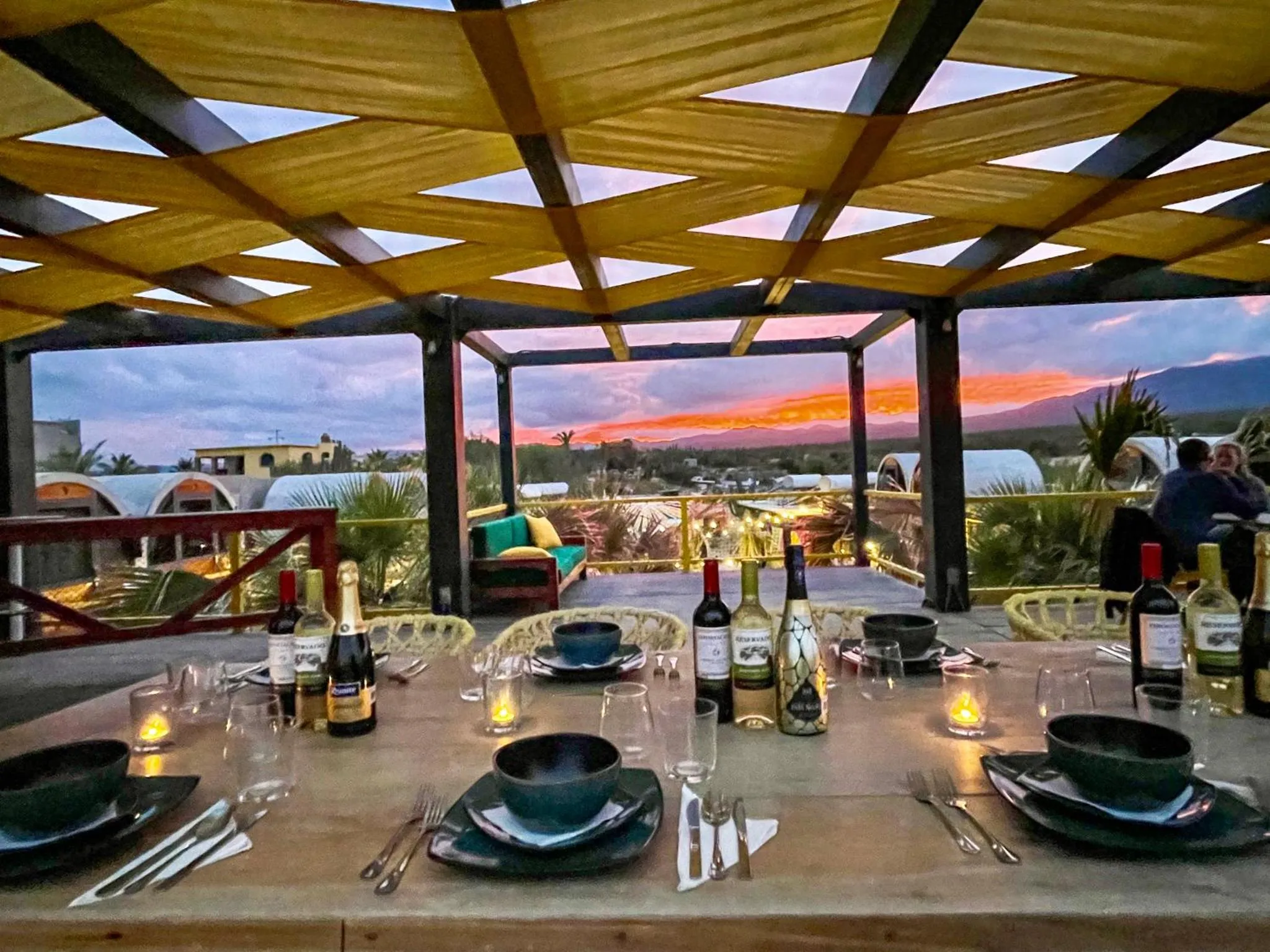Table dressée avec des bouteilles et des couverts sous une pergola ouverte sur un coucher de soleil.