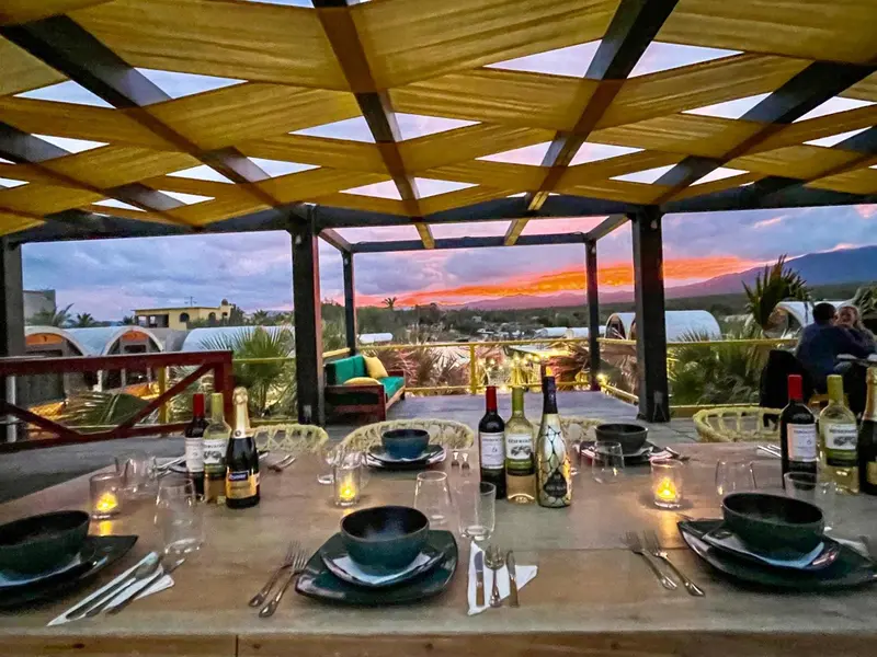 Table dressée avec des bouteilles et des couverts sous une pergola ouverte sur un coucher de soleil.