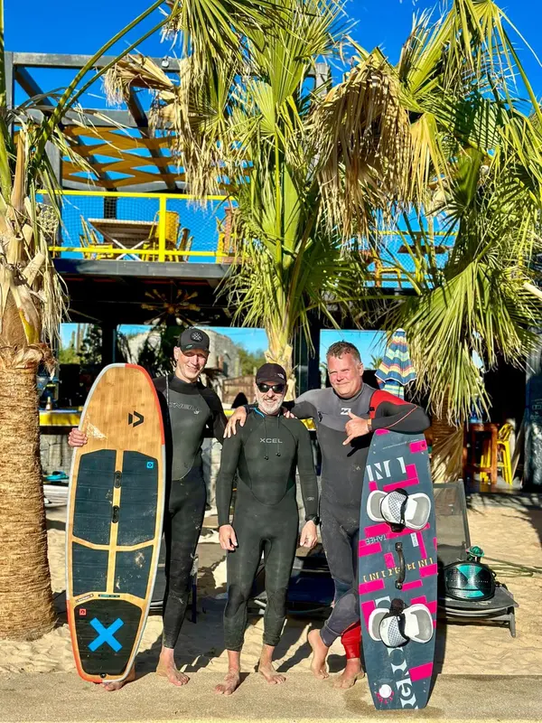 Trois personnes souriantes en combinaison de plongée se tiennent sur une plage ensoleillée.