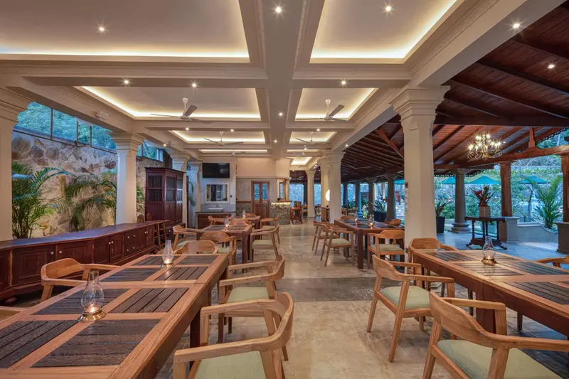 Une salle lumineuse avec des tables et des chaises en bois, éclairée par des luminaires encastrés et des ventilateurs de plafond.