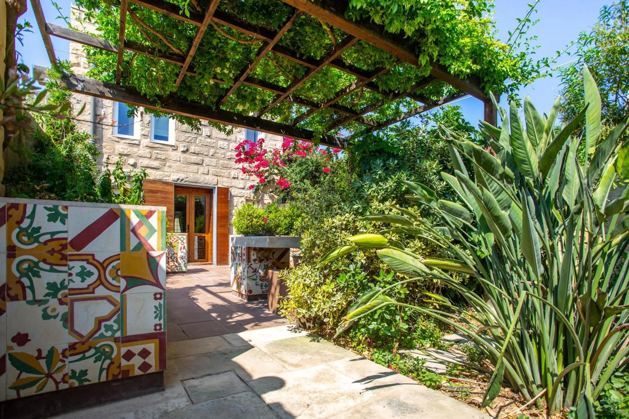 Une cour pavée ombragée par une pergola végétalisée et ornée de motifs colorés.