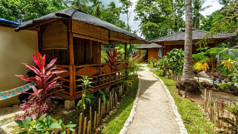 Des bungalows en bois sur pilotis bordent un chemin dans un jardin tropical.