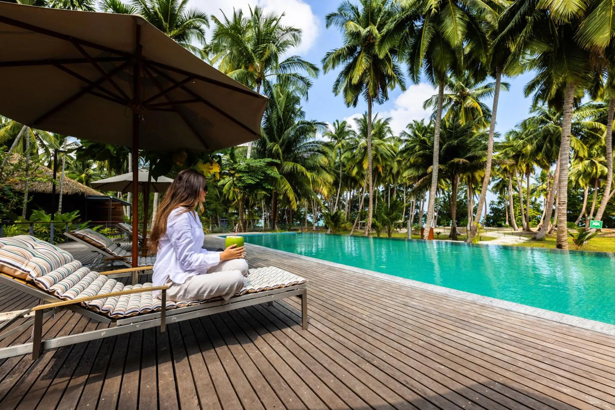 Une femme se détend sur un transat au bord d'une piscine turquoise ombragée par des parasols.
