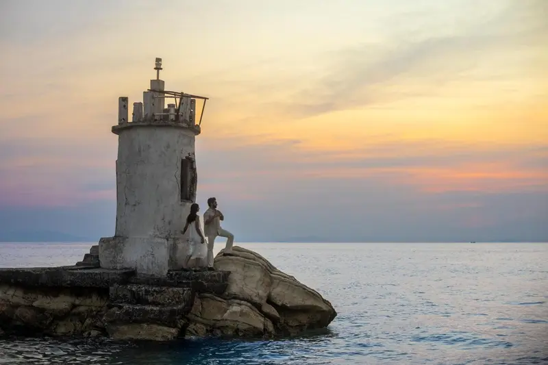Un phare blanc se dresse sur un promontoire rocheux au coucher du soleil.