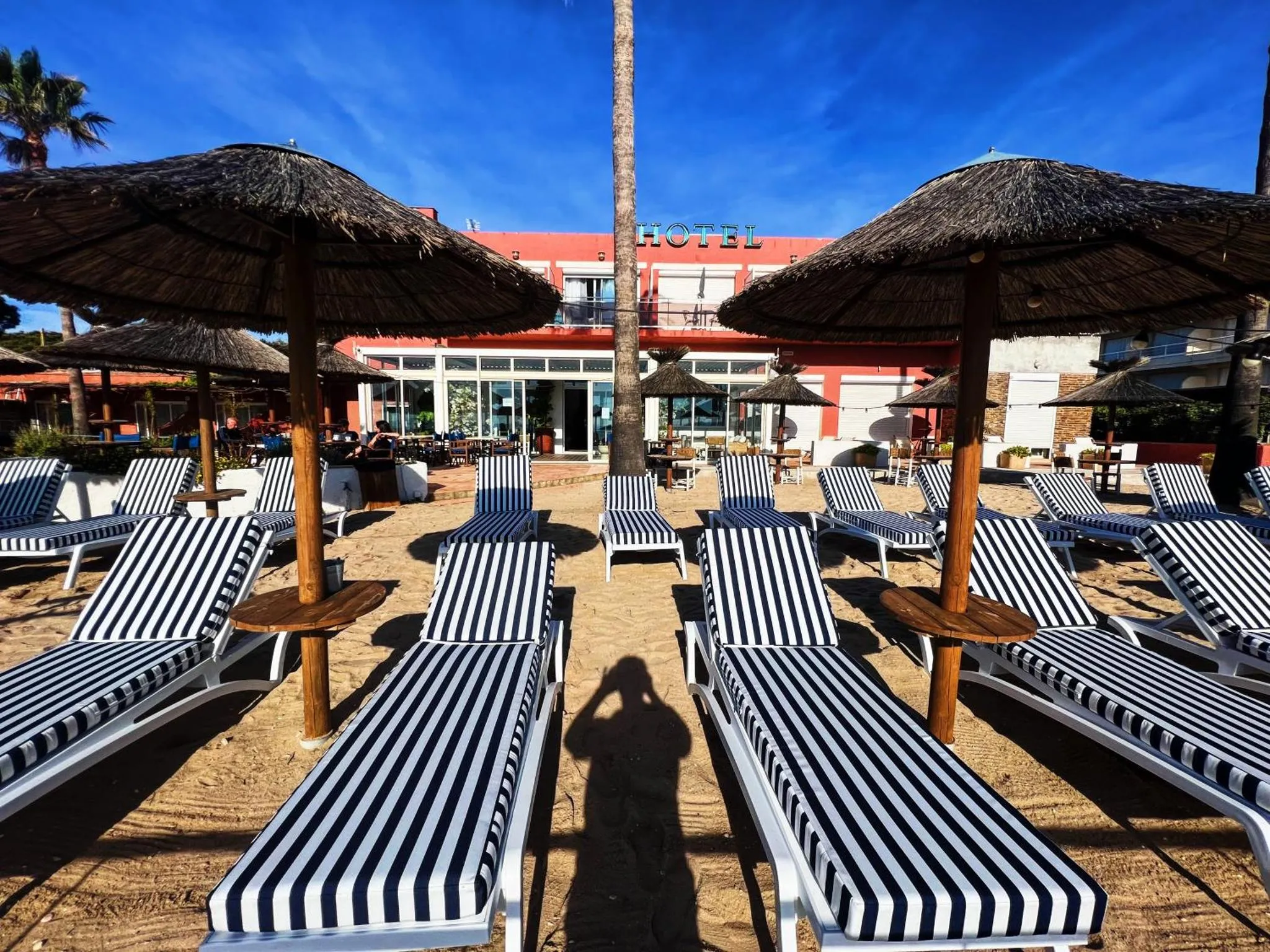 Transats rayés noirs et blancs alignés sur une plage de sable fin devant un bâtiment rouge.