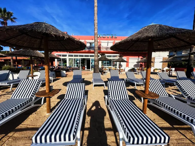 Transats rayés noirs et blancs alignés sur une plage de sable fin devant un bâtiment rouge.