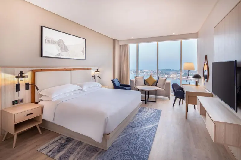 Chambre lumineuse avec un grand lit et un coin salon près d'une fenêtre.