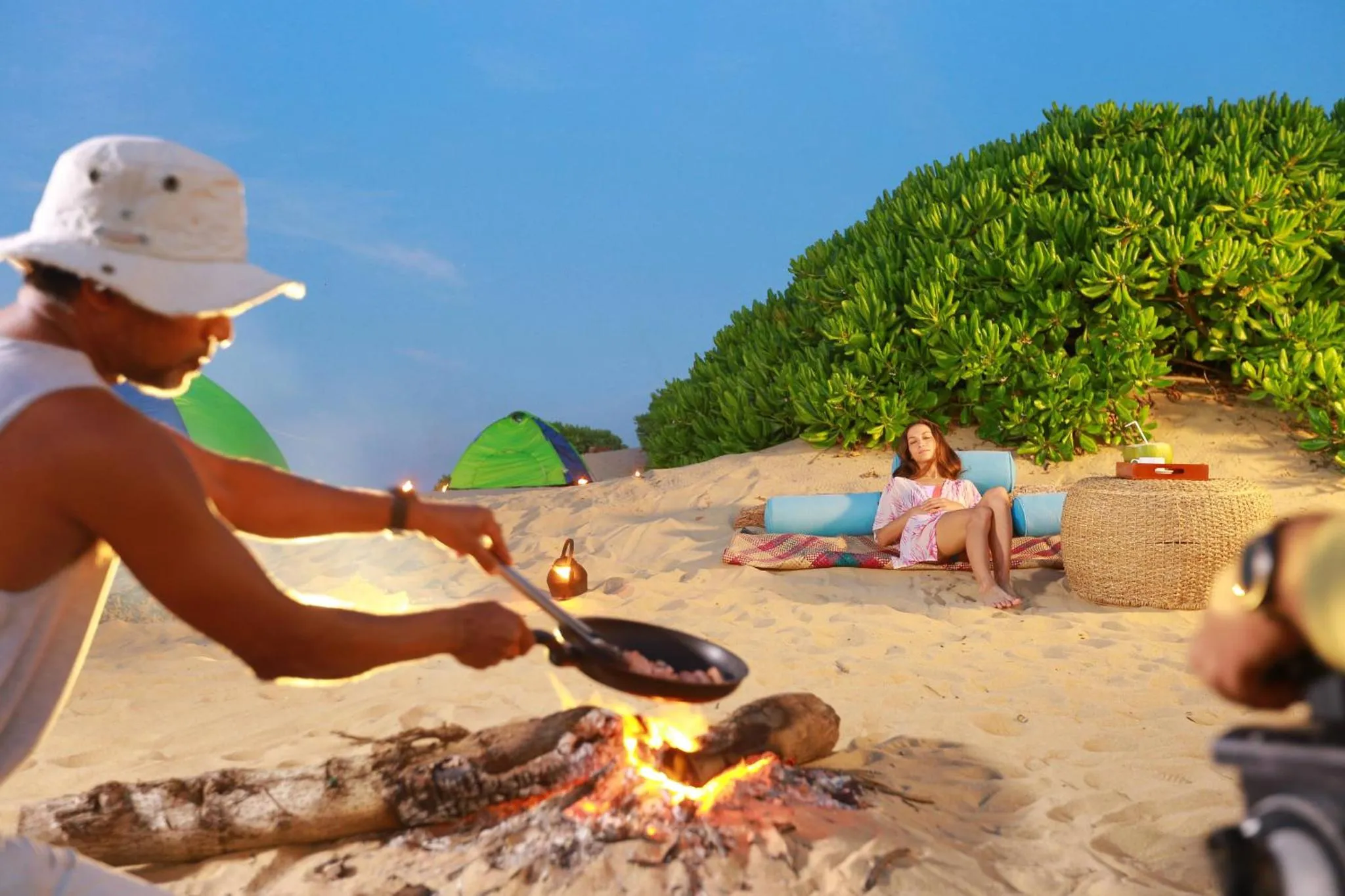 Un homme cuisine sur un feu de camp sur une plage de sable, tandis qu'une femme se détend sur un coussin.