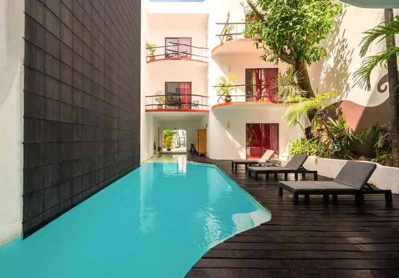 Une piscine turquoise bordée de chaises longues se trouve devant un bâtiment blanc et gris.