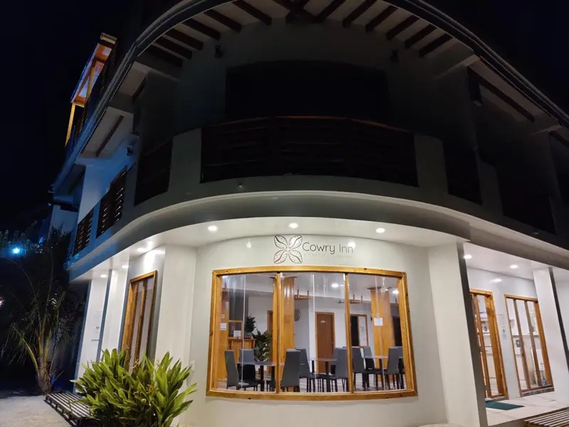 Façade d'un bâtiment blanc avec une entrée vitrée et une végétation tropicale.