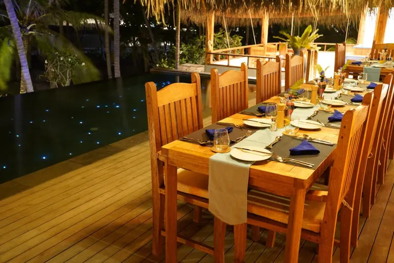 Une longue table en bois est dressée pour un dîner en extérieur, près d'une piscine éclairée.