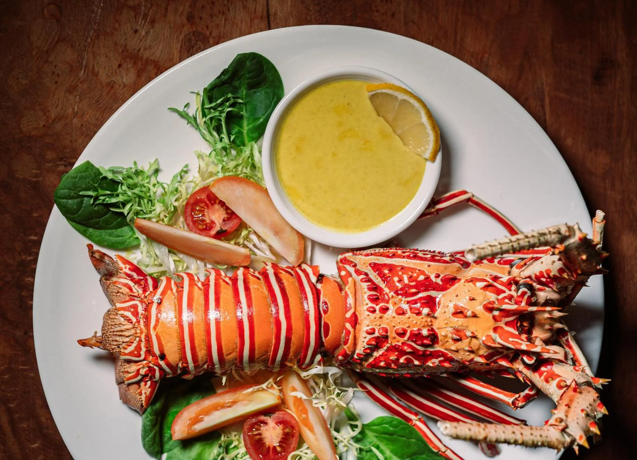 Un plat de homard rouge servi avec une sauce jaune, des légumes verts et des quartiers de citron.
