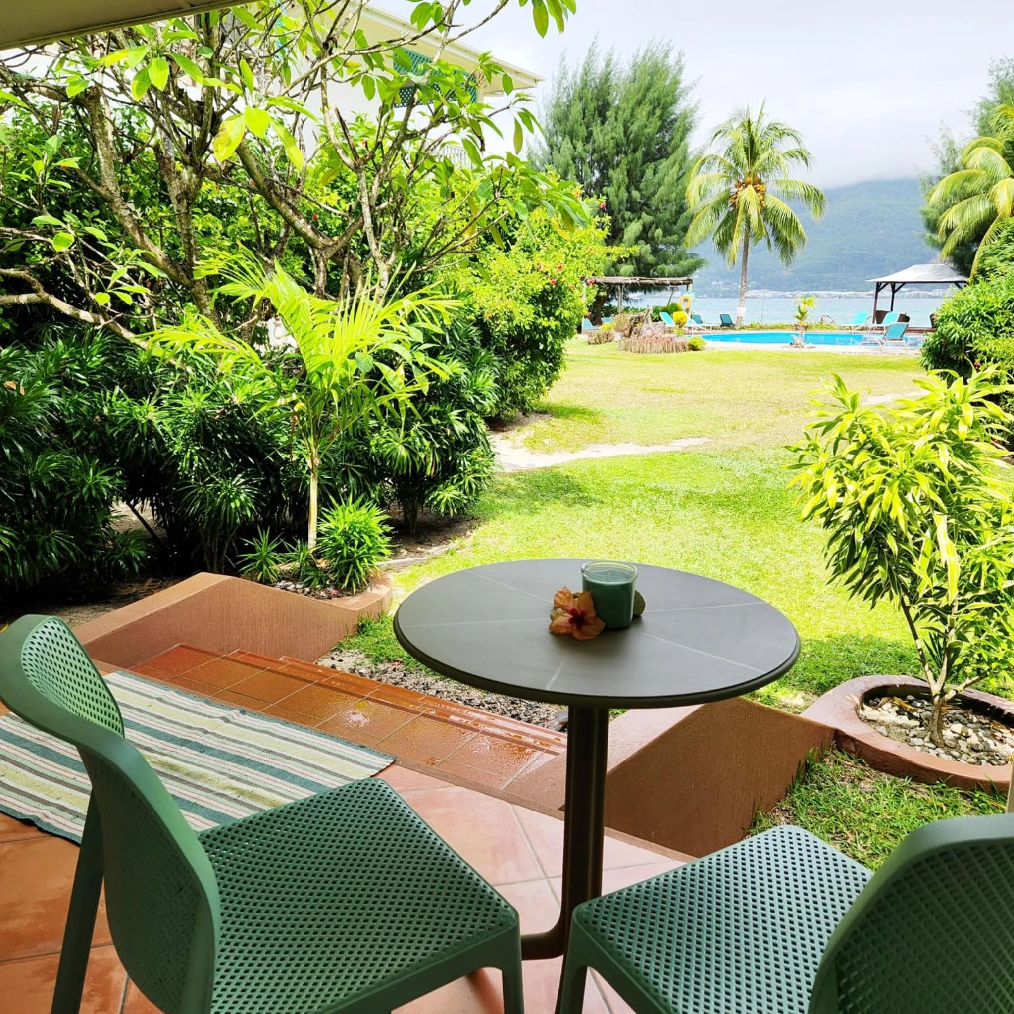 Une table et des chaises vertes se trouvent sur une terrasse ombragée avec vue sur un jardin et une piscine.