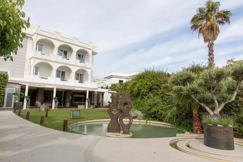 Façade blanche d'un bâtiment avec un jardin verdoyant et une fontaine au premier plan.