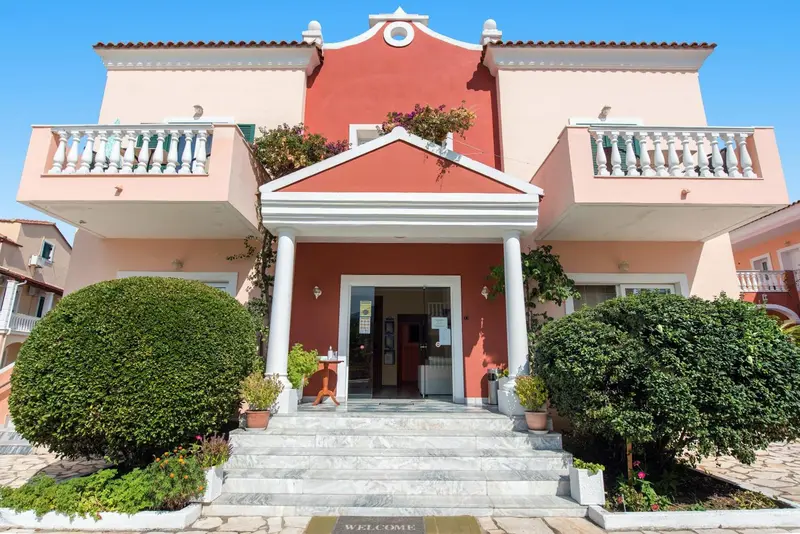 Facade d'un bâtiment rose et orange avec un escalier en pierre et des arbustes taillés.