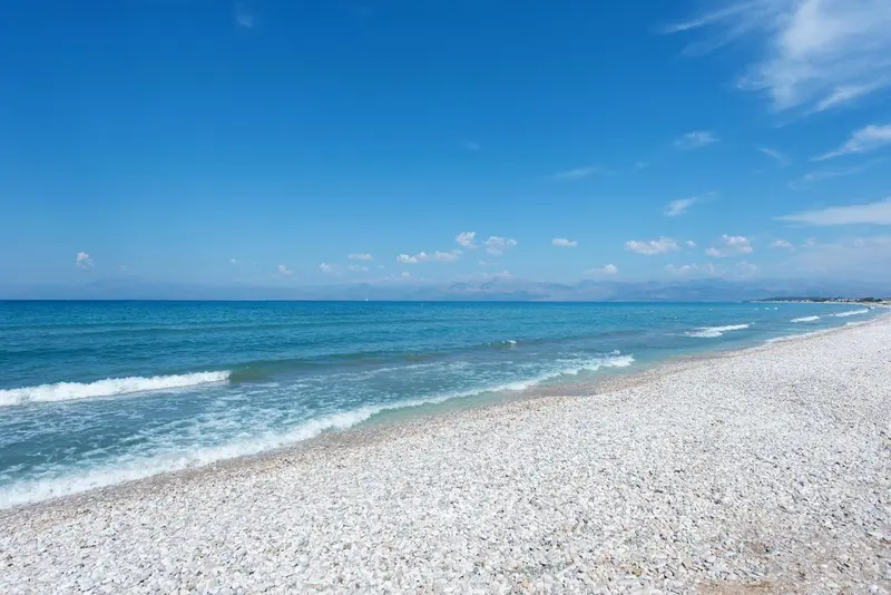 Une plage de galets blancs borde une mer turquoise sous un ciel bleu clair.