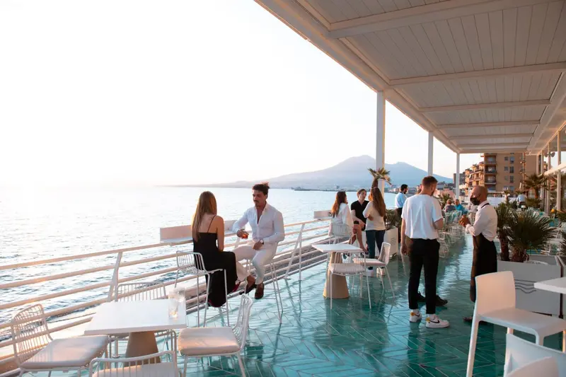 Des clients sont assis et discutent sur une terrasse ensoleillée avec vue sur la mer.