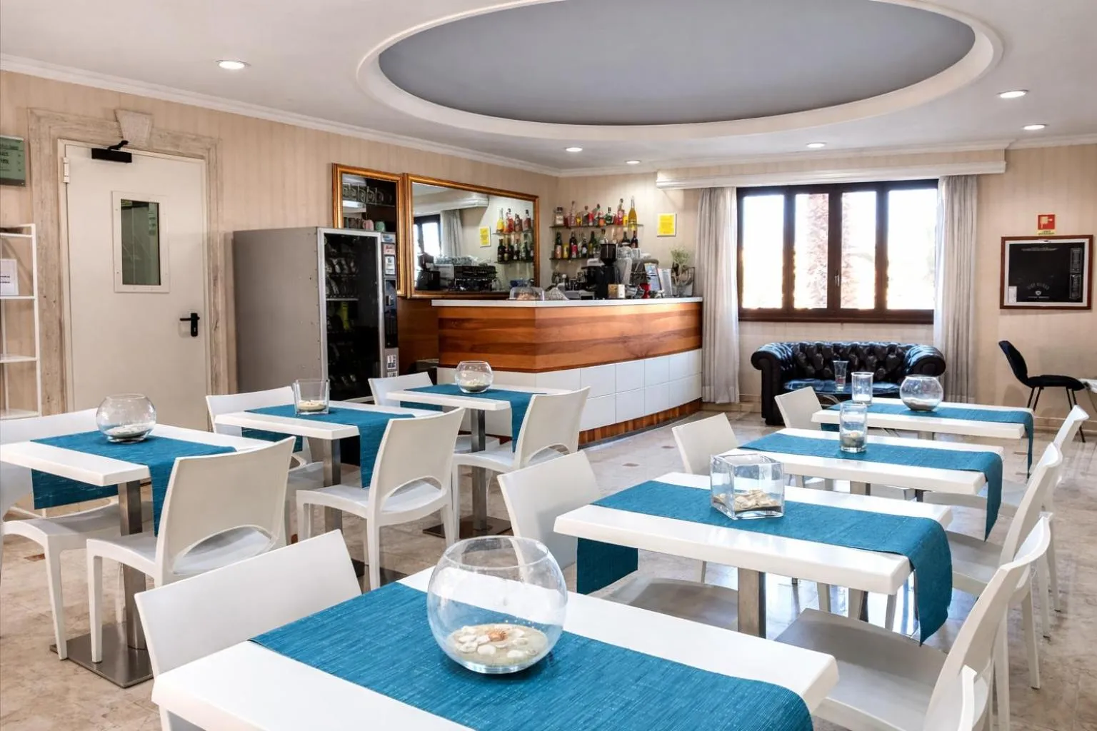 Une salle lumineuse avec des tables et des chaises blanches, un bar et un canapé.