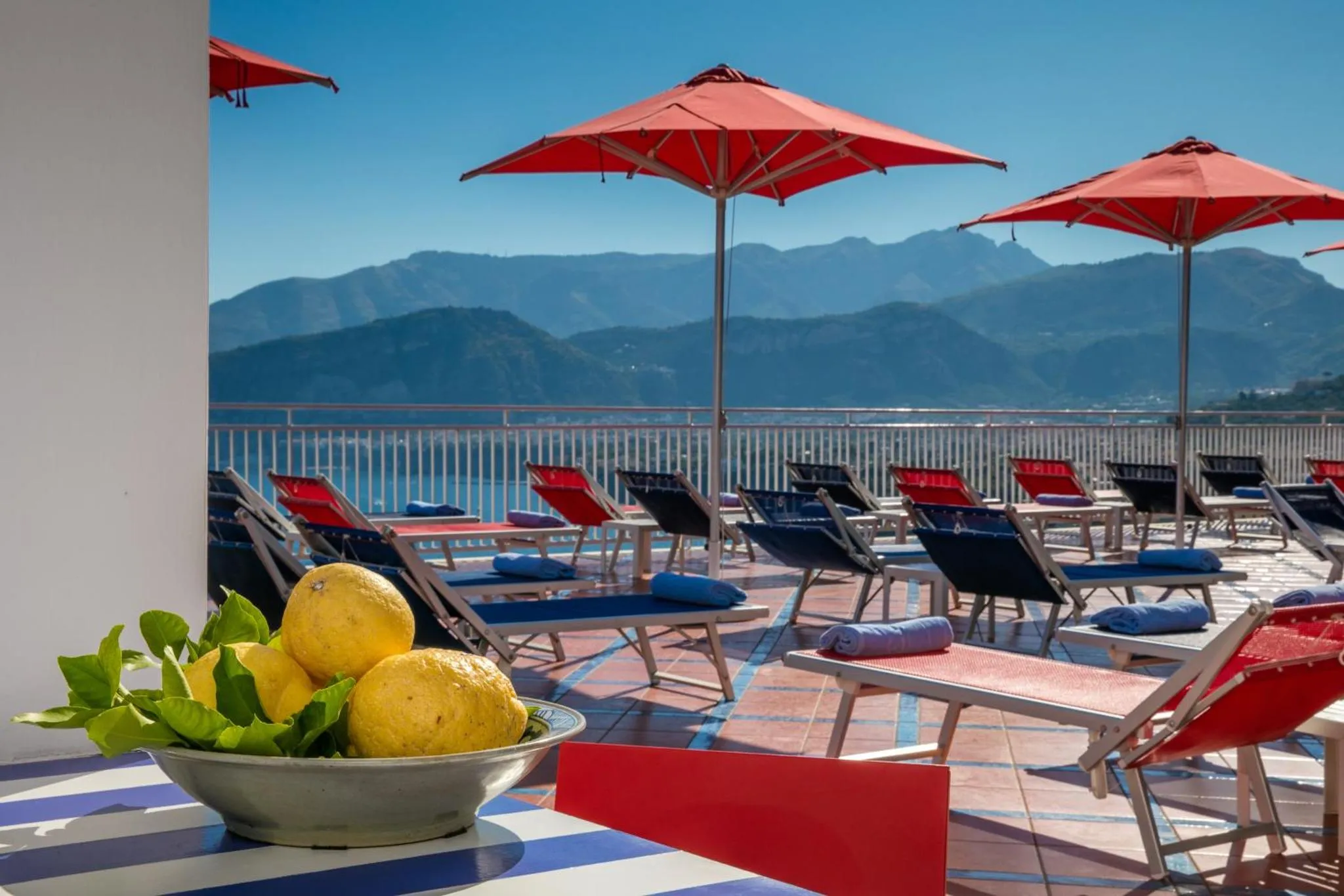 Transats rouges et bleus sous des parasols rouges, avec une vue sur la mer et les montagnes.