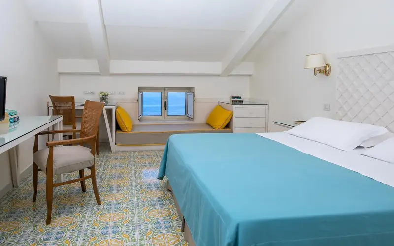 Une chambre lumineuse avec un lit recouvert d'une couverture turquoise et un coin lecture près d'une fenêtre.