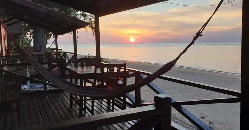 Un hamac est suspendu sur une terrasse en bois face à une plage au coucher du soleil.