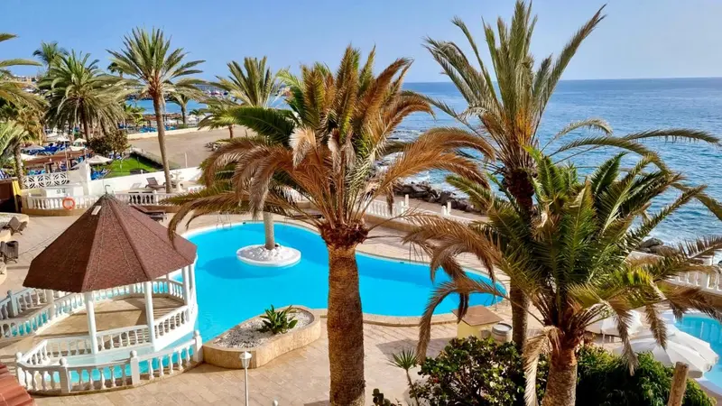 Une piscine turquoise bordée de palmiers et de transats, avec une vue sur la mer.