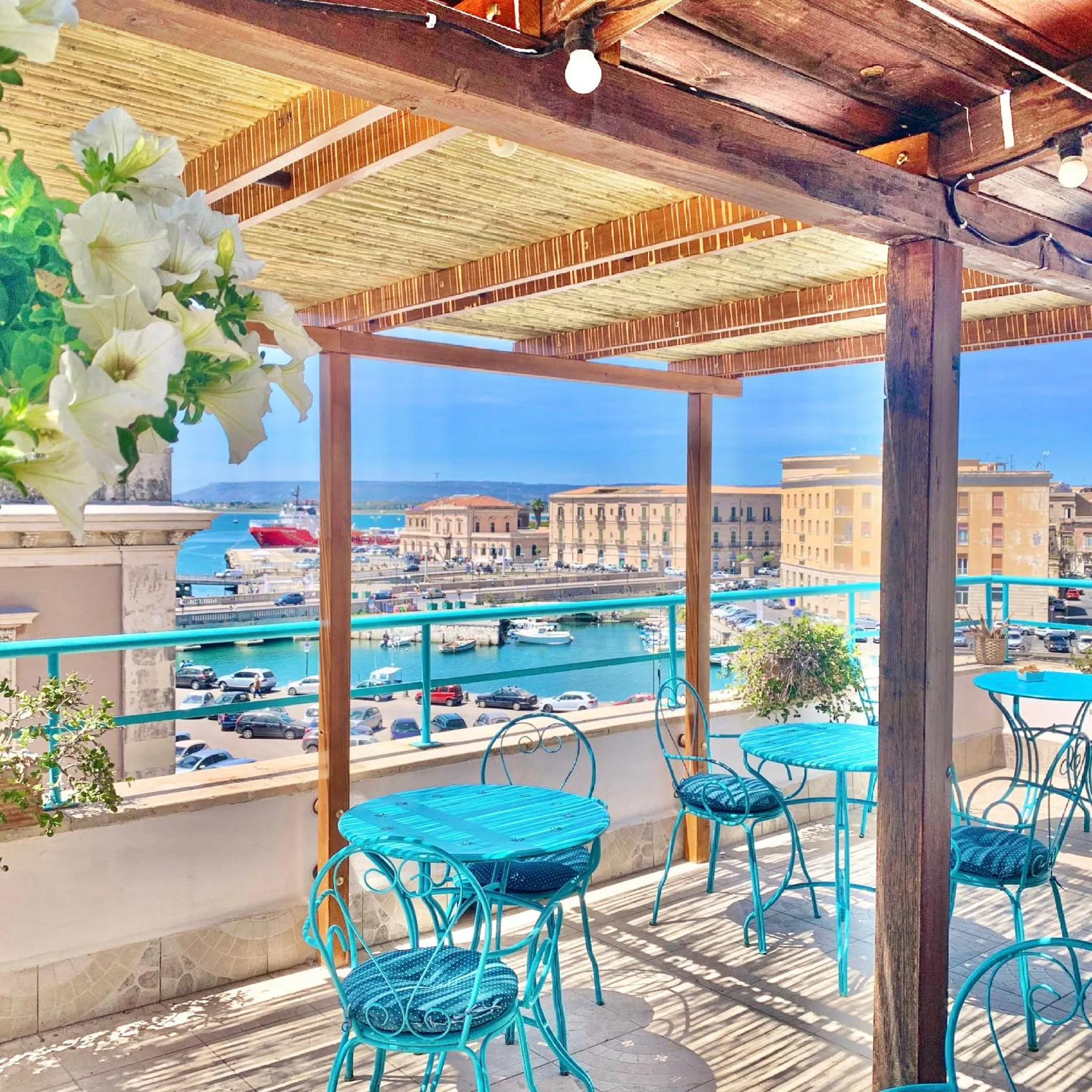 Terrasse ombragée avec tables et chaises donnant sur un port de ville.