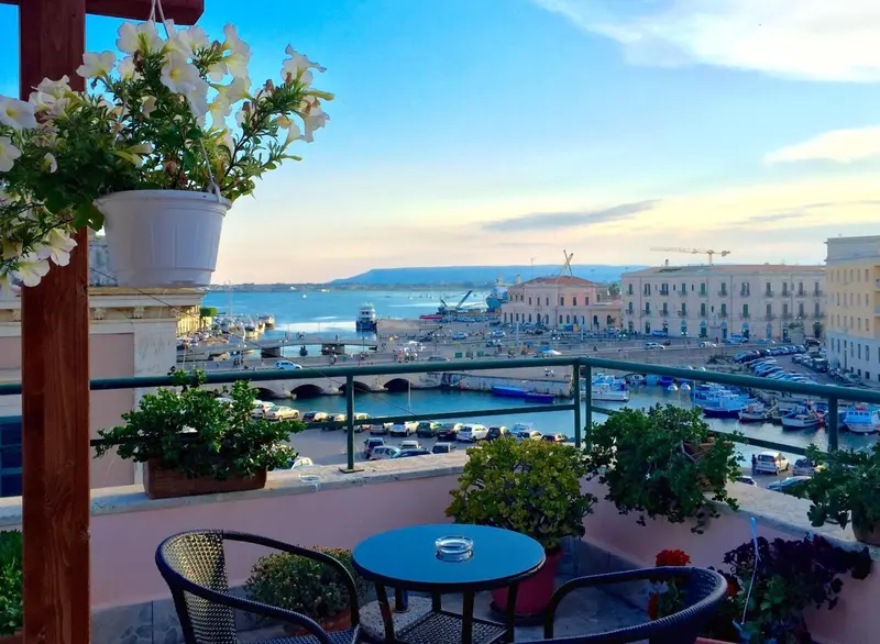Terrasse fleurie avec mobilier de jardin donnant sur un port et la mer.
