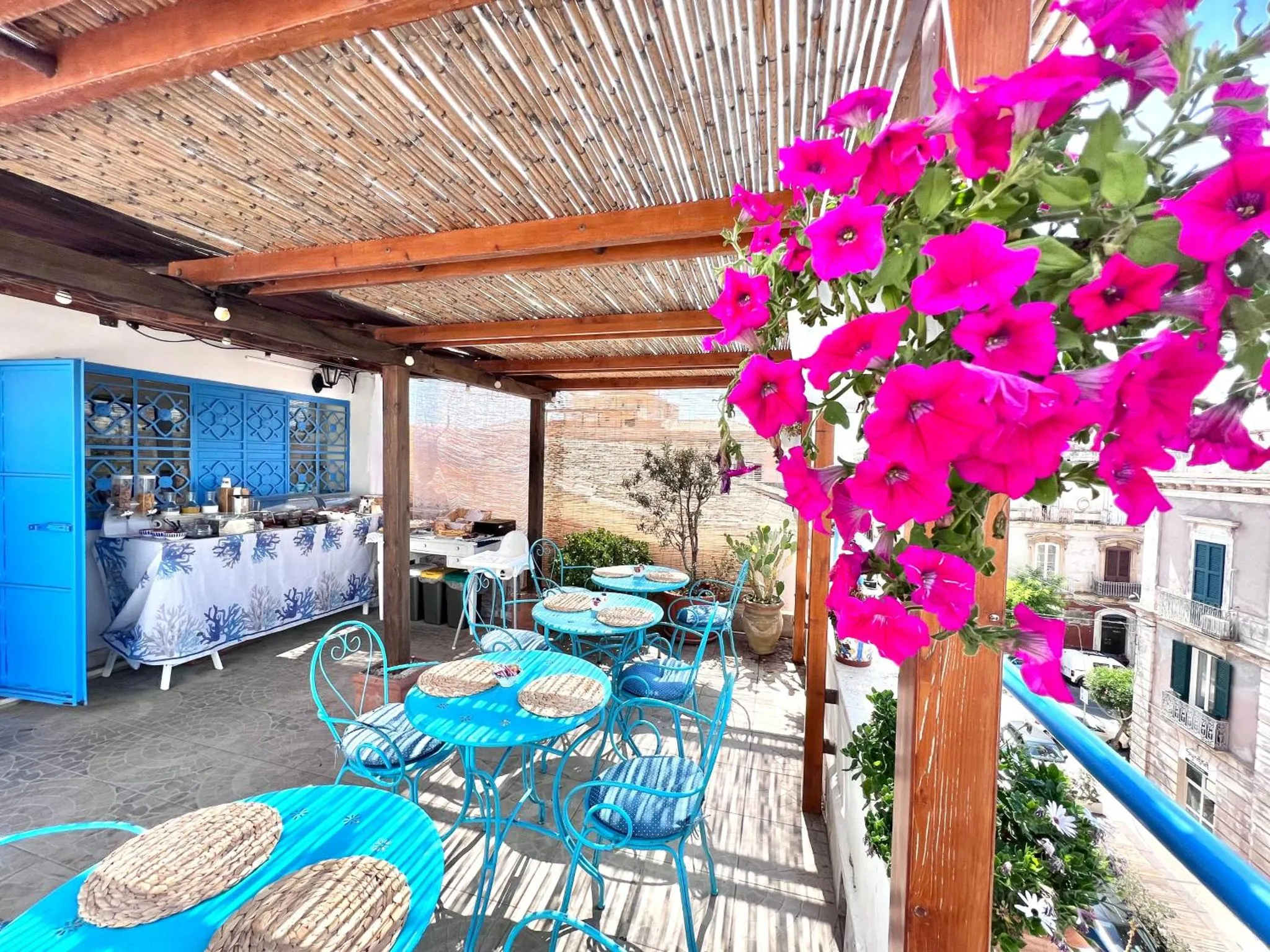 Terrasse ombragée avec tables et chaises bleues, agrémentée de fleurs roses et surplombant un paysage urbain.